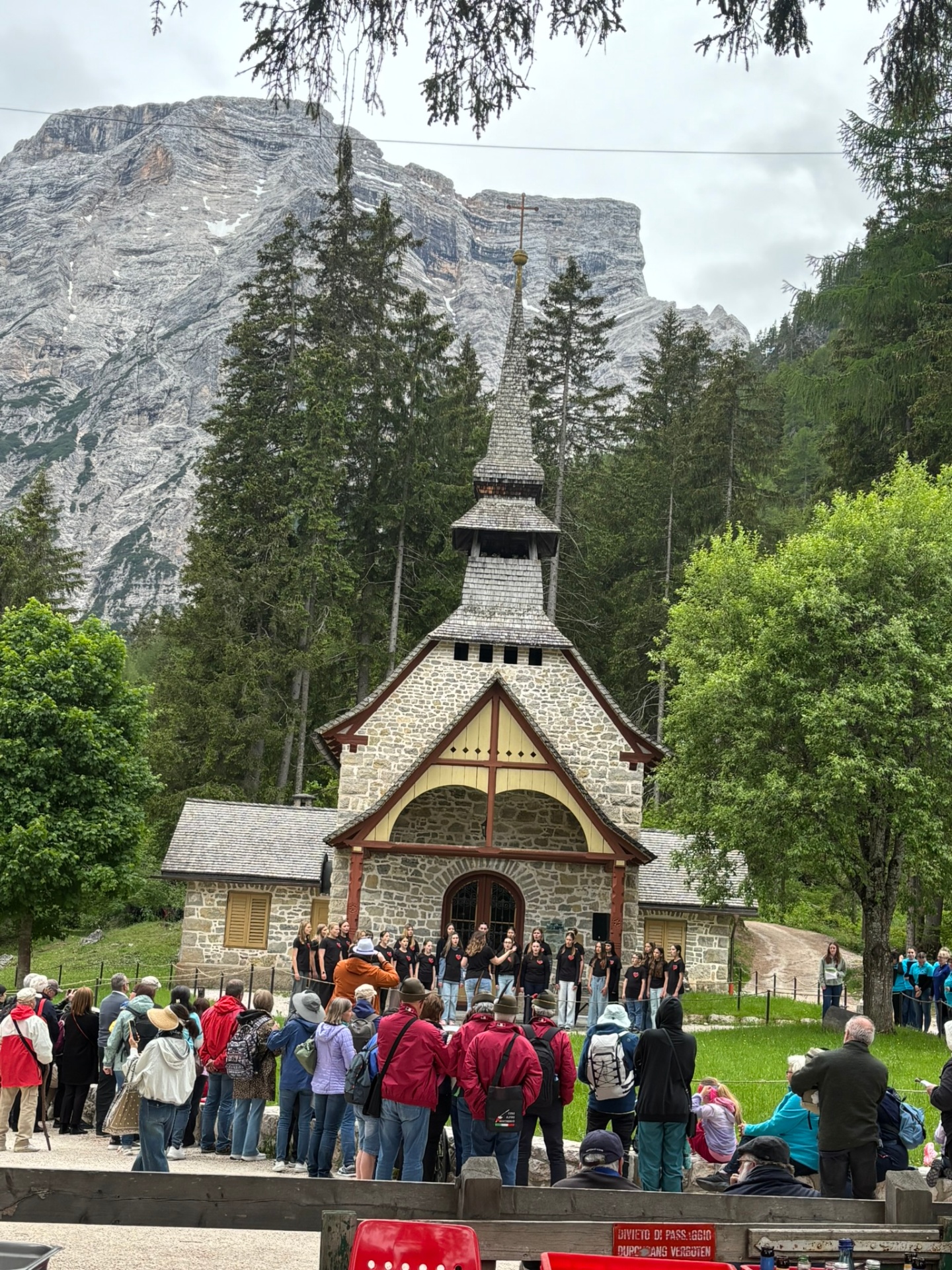 A youth choir in matching black shirts singing outside a small alpine chapel with the Dolomites rising behind them