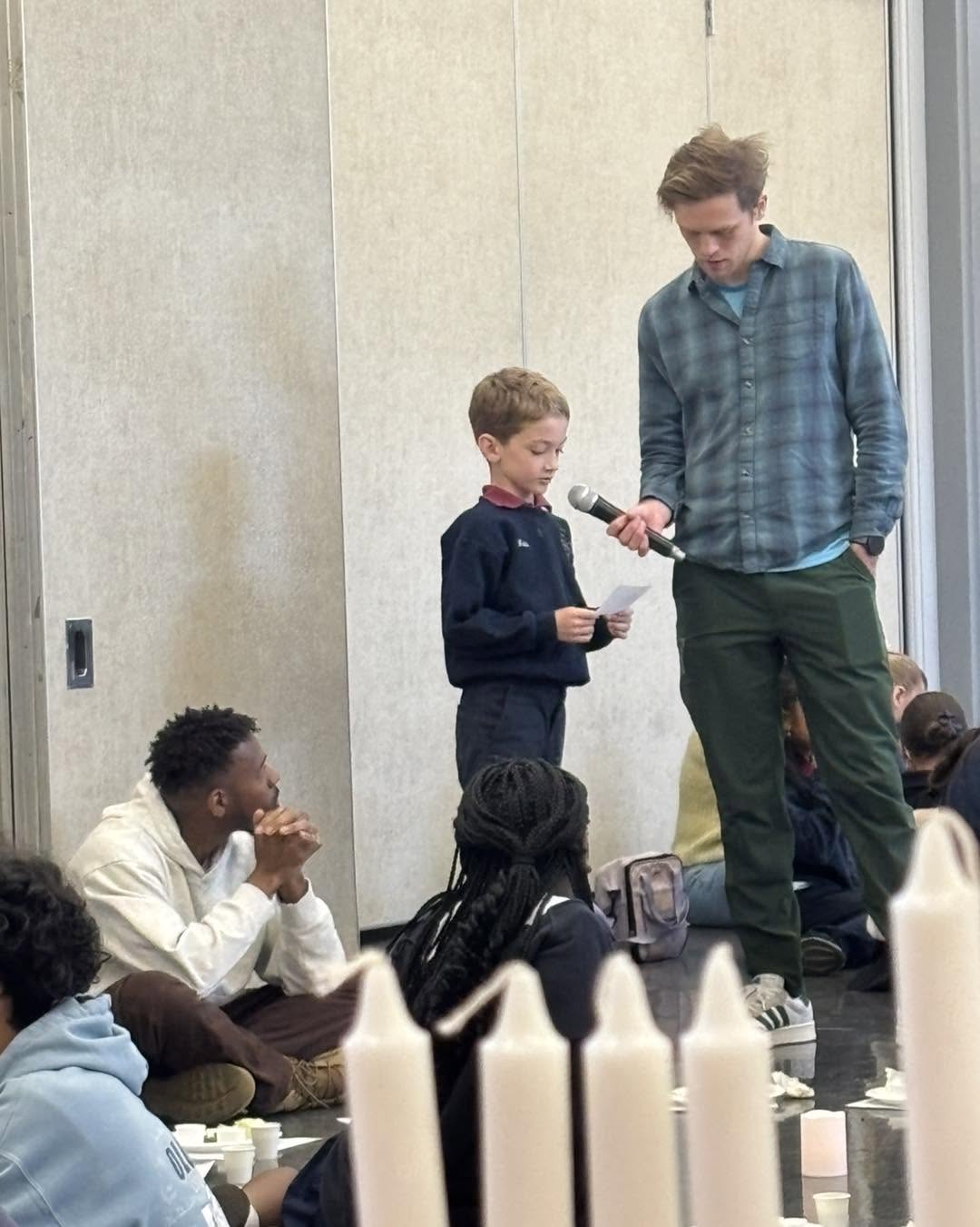 A teacher kneeling beside a student holding a microphone during a school gathering